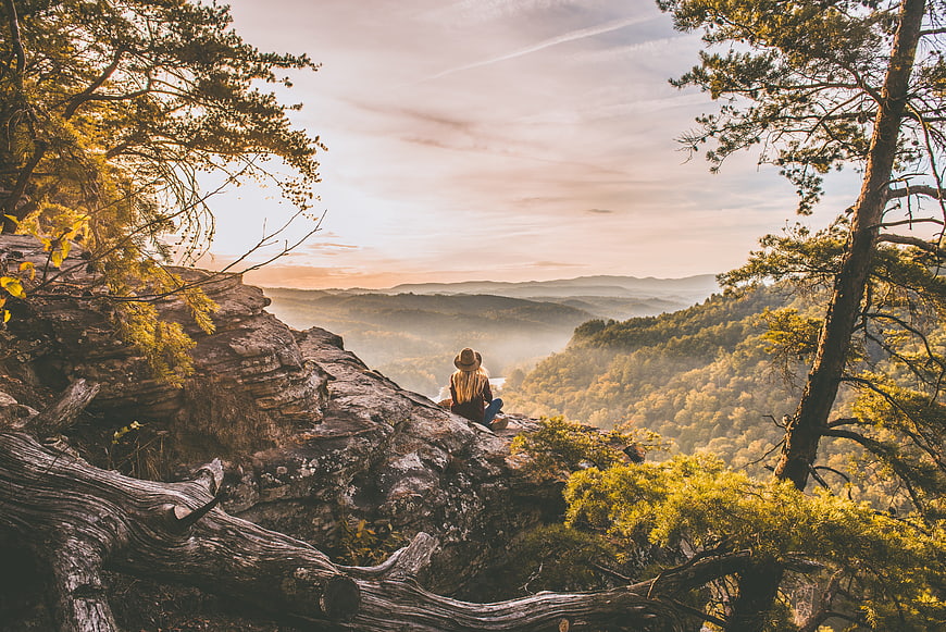 Woman sitting on rocky cliff overlooking misty mountain forest at sunrise