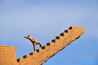 Sand sculpture of a figure climbing stairs against a blue sky