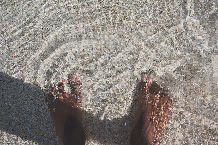 Bare feet in clear shallow water on pebbly beach