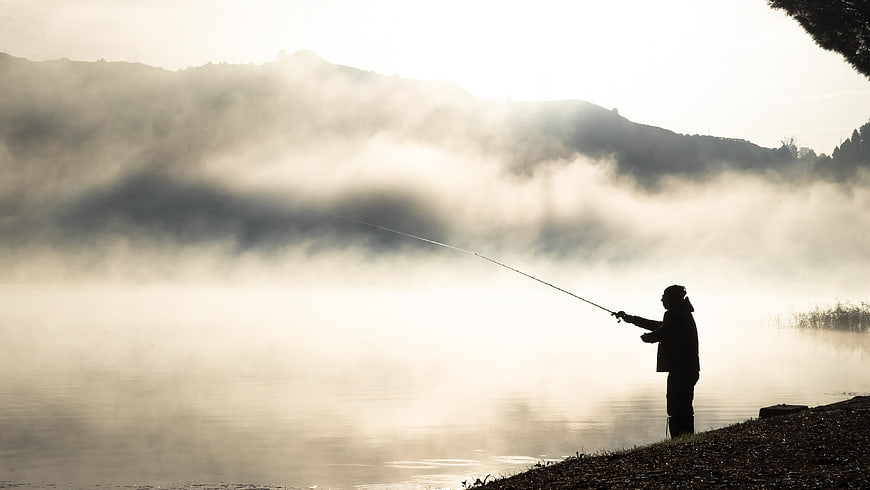 Silhouette of fisherman casting rod on misty lake morning