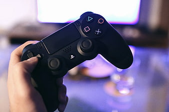 Hand holding black PlayStation controller with blurred screen background