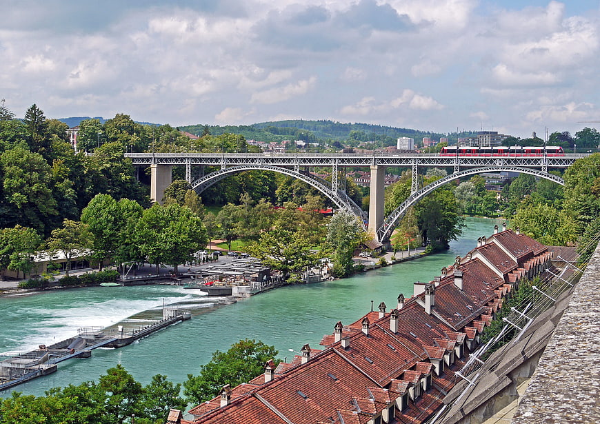 Red Train Crossing Bridge Over Turquoise River and Historic Roofs