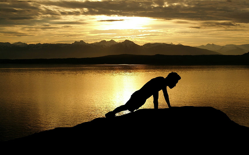 Silhouette of man doing push-up on rock by lake at sunset
