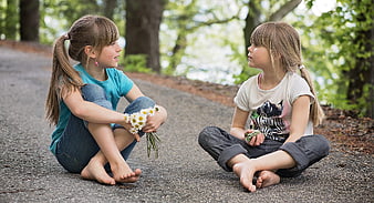 Two young girls sitting barefoot on a path talking with daisies
