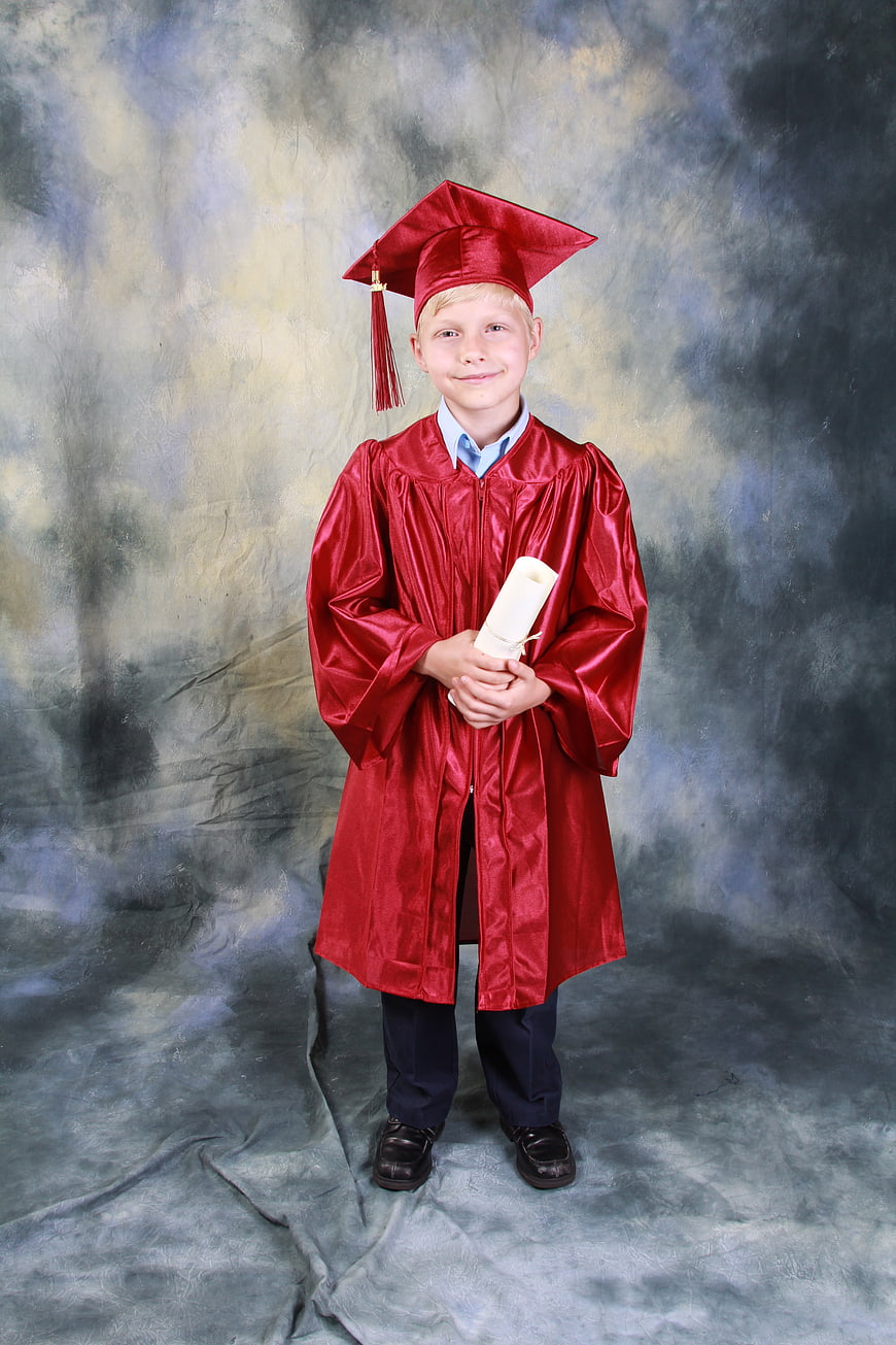 Young boy in red graduation gown holding diploma against studio backdrop