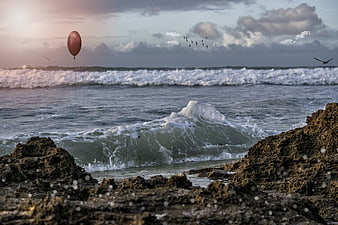 Red Balloon Floating Over Rocky Ocean Waves at Sunset