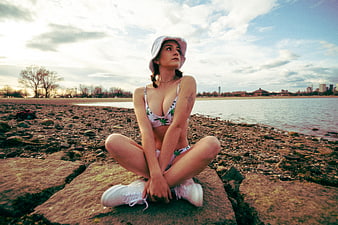 Young woman in floral bikini and bucket hat sitting on rocky shoreline