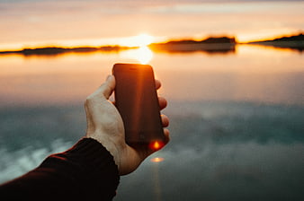 Hand holding smartphone capturing sunset over calm ocean water