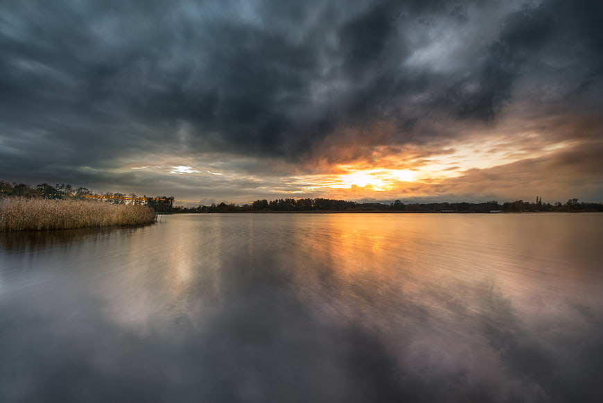Dramatic sunset over calm lake with reeds and storm clouds