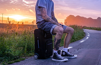 Tattooed Hitchhiker Sitting on Suitcase by Road at Sunset