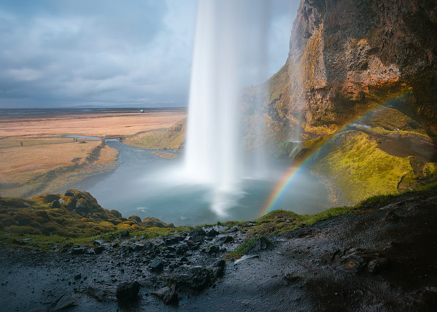 View from behind Skogafoss waterfall with rainbow and mossy rocks