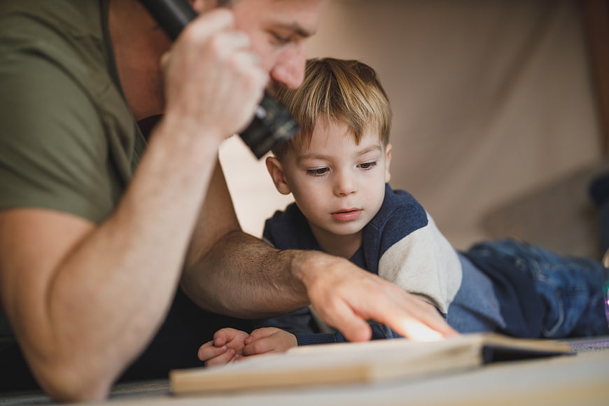 Father and son reading book with flashlight indoors