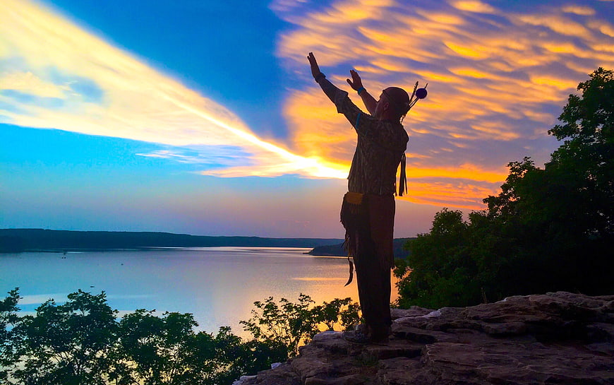 Man in Native American Attire Silhouetted Against Sunset Lake