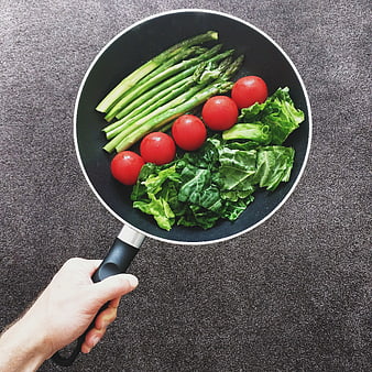 Hand holding frying pan with fresh asparagus, tomatoes, and kale