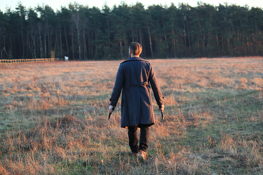 Man in Trench Coat Walking Through Field with Guns