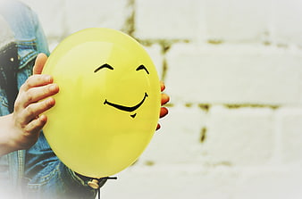 Child Holding Yellow Smiley Balloon Against White Brick Wall