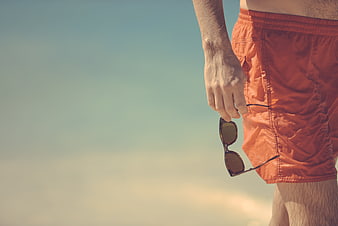 Man in orange swim trunks holding sunglasses against a blurred sky