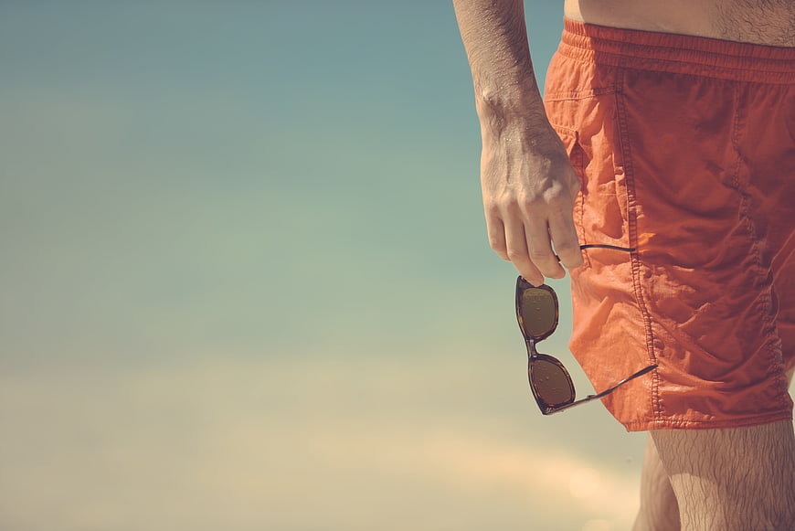 Man in orange swim trunks holding sunglasses against a blurred sky