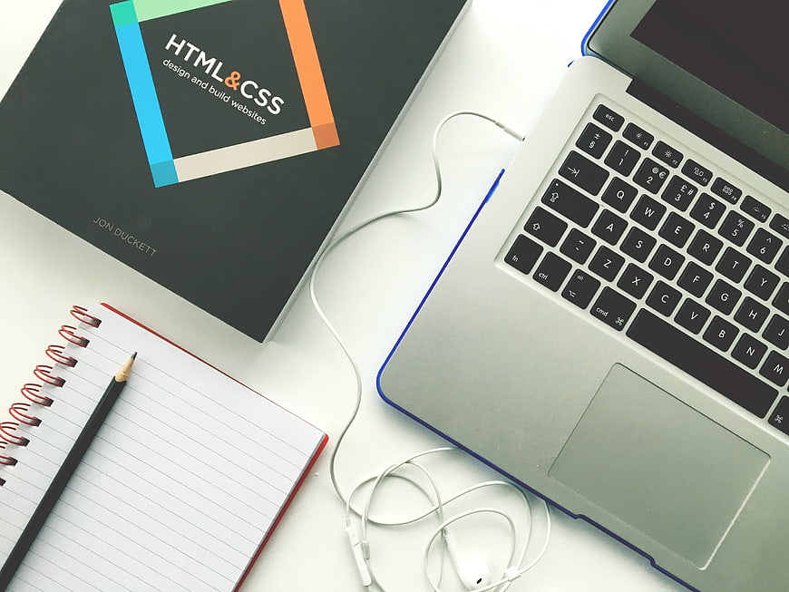 Flat lay of HTML CSS book, laptop, and notebook on white desk
