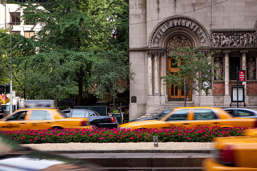 Yellow taxis speeding past ornate stone building with pink flowers