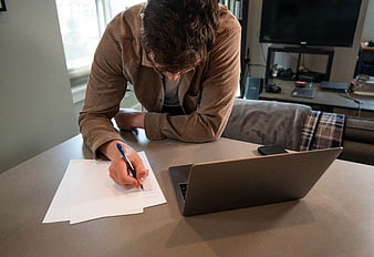 Man writing on paper at desk with laptop