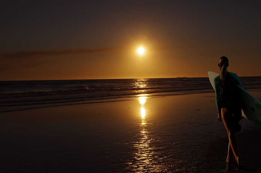 Woman with surfboard walking on beach at sunset
