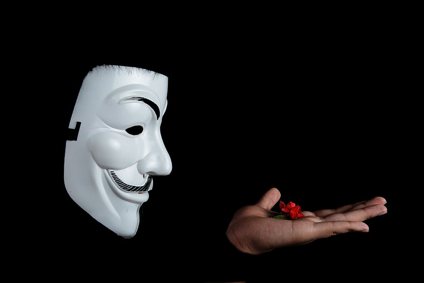Guy Fawkes mask and hand holding red flower on black background