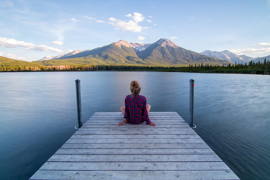 Woman Sitting on Wooden Dock Facing Serene Lake and Mountains