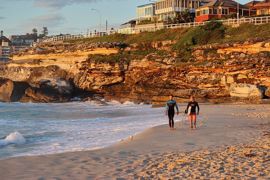 Surfers walking on sandy beach at sunset with cliffs
