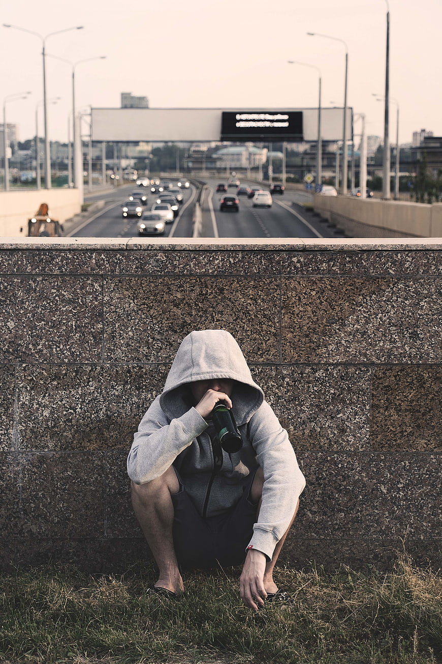 Lonely man in hoodie drinking from bottle against highway wall