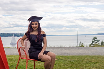Female graduate in black gown and cap sitting on red chair by lake