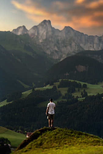 Man standing on grassy hill overlooking mountains at sunset