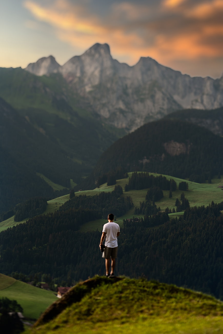Man standing on grassy hill overlooking mountains at sunset