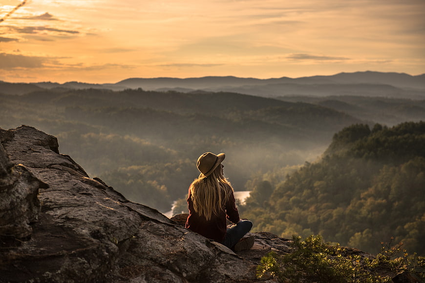 Woman in hat sitting on rocky mountain peak overlooking misty valley at sunset