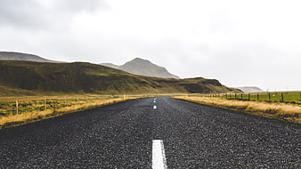 Empty asphalt road leading to green mountains under cloudy sky