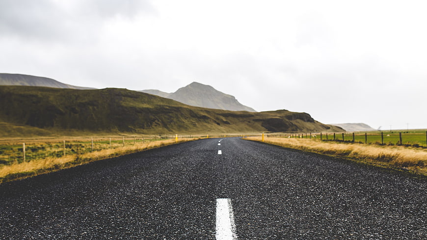 Empty asphalt road leading to green mountains under cloudy sky