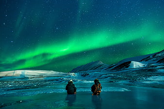 Two people watching vibrant green aurora borealis over snowy mountains