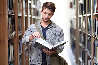 Young male student reading a book in a library aisle