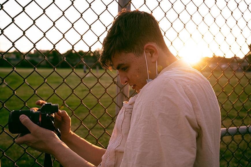Young Photographer Reviewing Camera by Chain Link Fence at Sunset