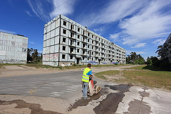 Worker operating plate compactor near abandoned concrete buildings