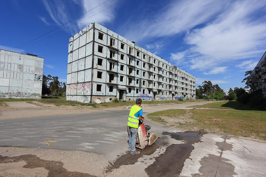 Worker operating plate compactor near abandoned concrete buildings