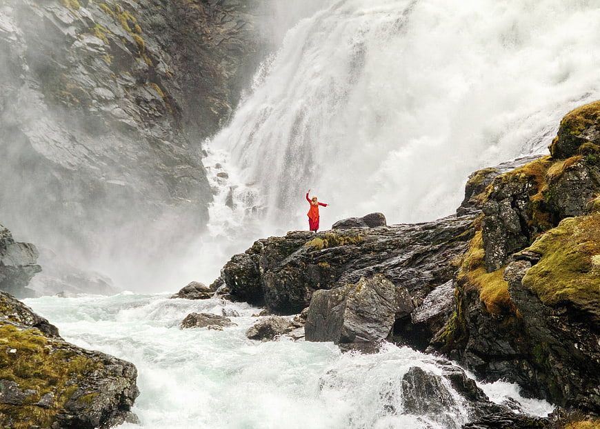 Woman in red dress standing on rocks in front of a large waterfall