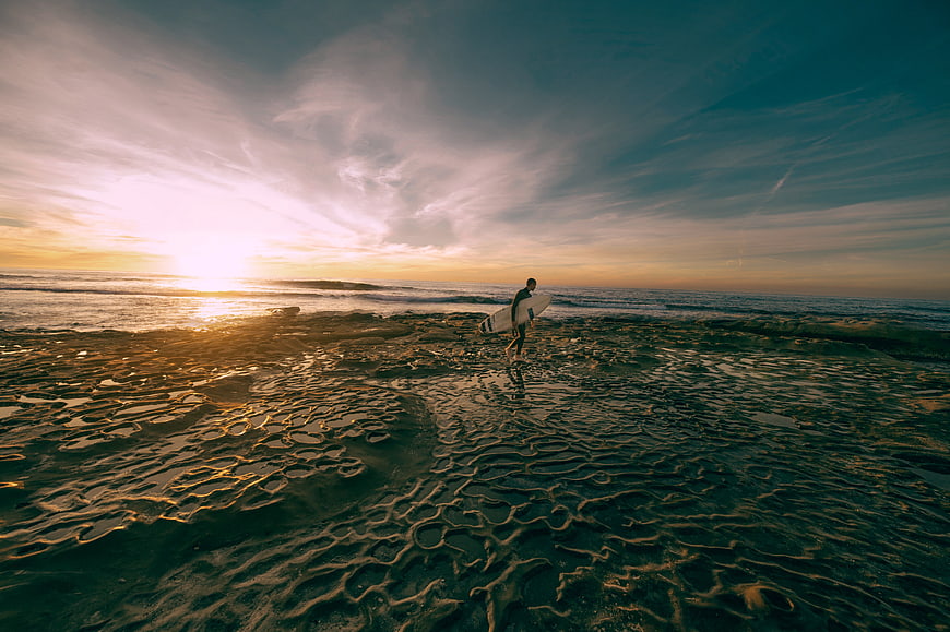 Surfer walking on wet sand beach at sunset with surfboard