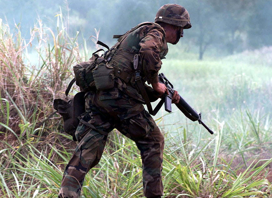 Soldier in camouflage gear moving through tall grass with rifle