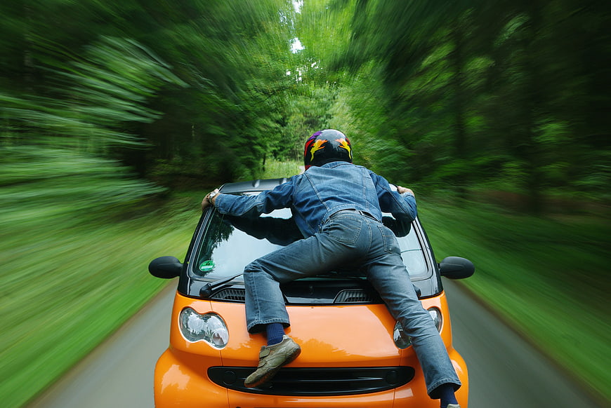 Man in helmet riding on hood of orange car speeding through forest