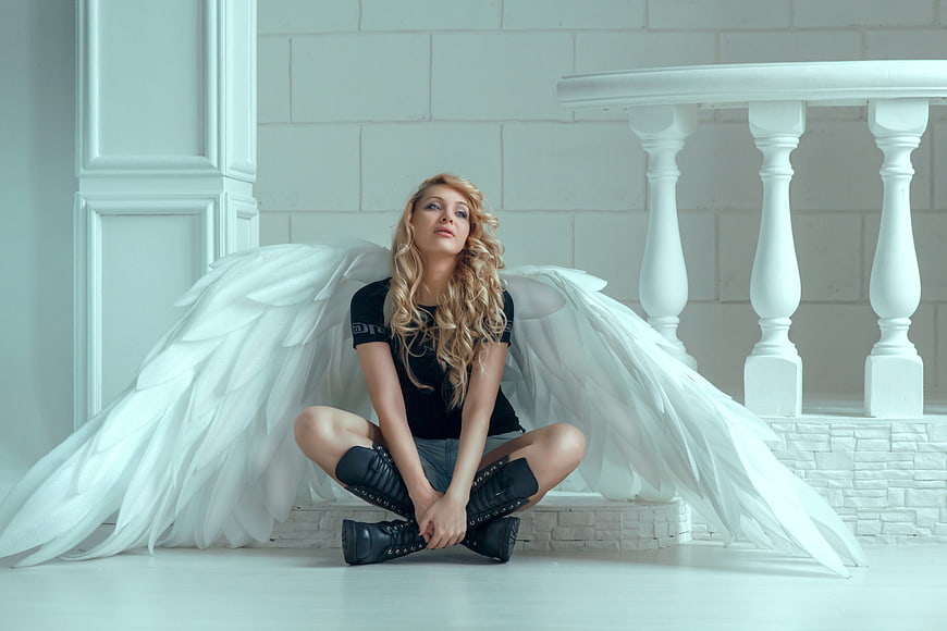 Blonde woman with white angel wings sitting in white room