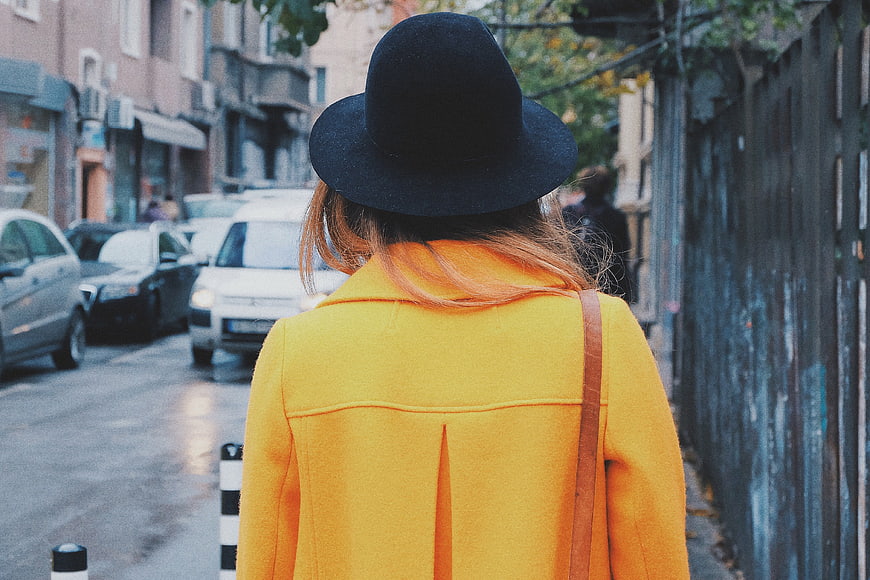 Woman in Yellow Coat and Black Hat Walking on City Street