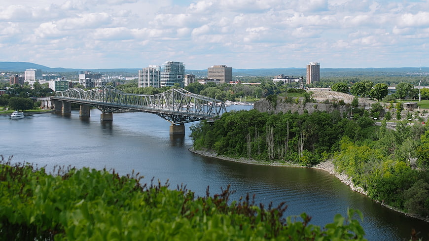 Steel truss bridge over river with city skyline and green cliffs
