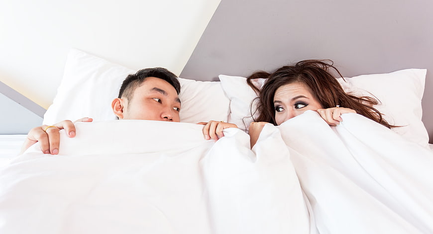 Asian couple peeking from under white duvet in bed