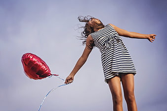 Woman holding red heart balloon against blue sky